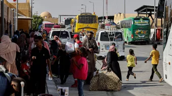 در تصویر، یک ایستگاه اتوبوس یا ترمینال مشاهده میشود که در آن تعدادی اتوبوس در حال توقف هستند. مردم زیادی در حال رفت و آمد هستند، برخی در حال حمل چمدان و بارهای خود هستند. در میان این افراد، کودکان نیز دیده میشوند که در حال بازی یا حرکت هستند. برخی از افراد در حال استراحت و نشستن بر روی ساکهای خود هستند. فضای ترمینال به نظر شلوغ و پرجنب و جوش میآید و آفتاب روشن در حال تابش است.