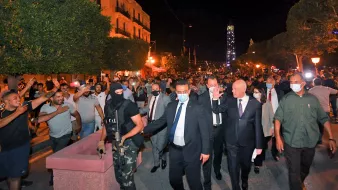 این تصویر گروهی از مردم را در یک خیابان در شب نشان میدهد که در حال راهپیمایی هستند. در میان آنها، یک فرد با لباس رسمی و گروهی از افراد دیگر حضور دارند. همچنین، یکی از افراد مسلح با یونیفورم مشخص در آنجا دیده میشود. به نظر میرسد که این صحنه به یک تجمع یا تظاهرات مرتبط باشد و تعدادی از معترضان و شخصیتهای دیگر در حال تعامل هستند. نورهایی از چراغهای خیابانی به وضوح در صحنه قابل مشاهده است.