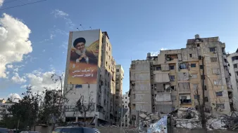 در این تصویر، یک ساختمان بزرگ با تصاویری از فردی که احتمالاً یک شخصیت عمومی است، دیده میشود. در کنار آن، ساختمانهای دیگری وجود دارند که به دلیل آسیبهای ناشی از زلزله یا جنگ، خرابه و آسیبدیده به نظر میرسند. وضعیت بناها نشاندهنده تخریب و ویرانی قابل توجهی است. آسمان در حال حاضر روشن است و ابرهای سفیدی در آن دیده میشود. سبزینگی در اطراف نیز وجود دارد که ممکن است درختان یا گیاهان باشد.