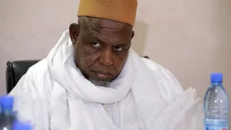 این تصویر یک مرد را نشان میدهد که لباسی سنتی به تن دارد و یک سرپوش قهوهای رنگ بر سر گذاشته است. او در حال نشسته و به جلو خیره شده است، به نظر میرسد که در حال تفکر یا گوش دادن به صحبتهای دیگران است. پسزمینه تصویری ساده و بدون هیچ جزئیات خاصی دارد.