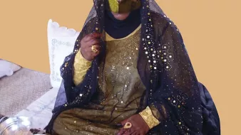 این تصویر یک زن را نشان میدهد که در لباس و پوشش سنتی خود نشسته است. او چهرهاش را با نوار مشکی پوشانده و چشمهایش نمایان است. لباس او شامل رنگهای تیره و زینتهای طلایی است که درخشندگی خاصی دارد. زن در حال نشستن بر روی یک سجاده یا پارچه با خطوط افقی است و در کنار او چند وسیله مانند ظرفهای کوچک دیده میشود. زمینه تصویر نیز به رنگ قهوهای ملایم است و به عکس حس گرم و جذابی میدهد.