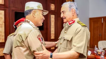 در این تصویر، دو شخص نظامی در حال گفتگو و تبادل نظر هستند. یکی از آنها Uniform نظامی با رنگهای بیابانی و شَمشیر نشاندار پوشیده است و دیگری یونیفرم نظامی با رنگهای مشابه دارد. فضای محیط رسمی و دارای دکور چوبی است. به نظر میرسد که این دو نفر در یک جلسه یا دیدار رسمی حضور دارند و بین آنها احترامی دوستانه و صمیمی برقرار است.
