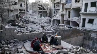 تصویر نشاندهندهی یک منطقه ویران در یک شهر است که آثار تخریب شدید در آن مشهود است. ساختمانها به شدت آسیب دیده و دیوارها و سقفها خراب شدهاند. در وسط این ویرانی، گروهی از افراد نشستهاند و روی زمین، میان خرابهها، در حال بازی یا سرگرمی هستند. فضای کلی تصویر حس ناامیدی و خرابی را منتقل میکند، اما در عین حال حضور انسانها نشانهای از امید و زندگی در شرایط سخت است.