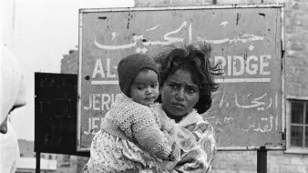 این تصویر یک کودک را نشان میدهد که در آغوش یک زن ایستاده است. زن ظاهراً نگران و جدی به نظر میرسد و کودک در آغوشش پایش را به دور او حلقه کرده است. پسزمینه تصویر شامل یک تابلو با نوشتهها به زبان عربی و انگلیسی است که نام مکانی را نشان میدهد. حالت چهره و لباسهای آنها نشاندهنده سختیهای احتمالی در زندگی است. تصویر به وضوح وضعیت انسانی و احساسات عمیق را منتقل میکند.