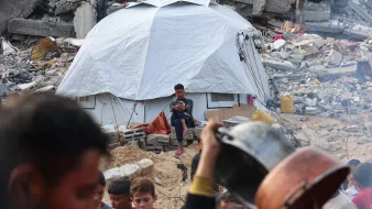 در این تصویر، یک چادر سفید در میان خرابهها قرار دارد و یک نوجوان در حال نشستن و استفاده از گوشی تلفن همراه است. در جلوی چادر، یک گروه از کودکان و افراد بزرگسال دیده میشوند که به سمت دوربین نگاه میکنند یا در حال انجام کارهایی هستند. زبالهها و خرابههای ساختمانهای تخریبشده در پسزمینه به وضوح احساس ناامیدی و بحران را منتقل میکنند.