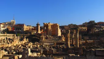 این تصویر نمایی از یک مکان باستانی را نشان میدهد که شامل خرابههای ستونها و دیوارهای قدیمی است. در پسزمینه، چند ساختمان مدرنتر دیده میشود که نشاندهنده ترکیبی از تاریخ با معماری معاصر است. آسمان آبی و صاف است و نور خورشید بر روی سنگها میتابد که حس زیبایی و آرامش را به تصویر میبخشد. اجزای باستانی این مکان نشاندهنده تاریخ غنی و فرهنگی آنجا هستند.