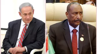 این تصویر دو شخصیت سیاسی را نشان میدهد. در سمت چپ، مردی با موهای خاکستری و لباسی رسمی به رنگ سیاه و با کراواتی رد و سفید دیده میشود. او در حالتی فکرکرده و آرام نشسته است. در سمت راست، مرد دیگری با صورتی جدی و لباسی رسمی به رنگ سیاه و نشان کشوری بر روی جیب خود نشسته است. این دو نفر احتمالاً در یک نشست یا کنفرانس در حال گفتگو هستند. در پسزمینه، تعدادی افراد دیگر مشاهده میشوند.