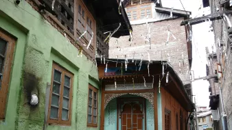 این تصویر نشاندهنده یک خیابان باریک در یک منطقه قدیمی است. ساختمانها به سبک سنتی ساخته شدهاند و رنگهای مختلفی دارند. دیوار یک ساختمان به رنگ سبز و دیگری به رنگ نارنجی است که با جزئیات چوبی تزئین شده است. درب چوبی و پنجرههای با قاب چوبی، زیبایی خاصی به این فضا بخشیدهاند. درختان و لامپهای آویزان نیز جزو دیگر عناصر موجود در تصویر هستند که به فضایی دلانگیز و سنتی افزودهاند.