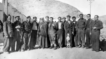 در این تصویر گروهی از مردان در یک محیط بیرونی تجمع کردهاند. آنها به طور رسمی لباس پوشیدهاند و در کنار هم ایستادهاند. در میان آنها یک مرد با لباس سنتی و یک سگ دیده میشود. پسزمینه تصویر شامل تپههای خشک و ساختمانهایی به نظر میرسد که ویژگیهای یک منطقه کوهستانی را نشان میدهد. نور تصویر قدیمی به نظر میرسد و به حالت سیاه و سفید است که به اثر حس نوستالژی خاصی میبخشد.