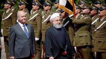 در این تصویر، دو شخصیت در حال برگزاری یک مراسم رسمی هستند. در سمت چپ، فردی با لباس رسمی و کت و شلوار ایستاده است. در سمت راست، فرد دیگری با لباس سنتی ایرانی، که به یک عمامه و عبای سیاه پوشیده است، حضور دارد. در پسزمینه، تعدادی از نظامیان لباسپوشیده به خط ایستادهاند و در حال انجام تشریفات نظامی هستند. تصویر نشاندهنده یک رویداد رسمی یا دیدار دیپلماتیک است.