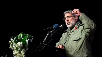 در این تصویر، یک فرد در حال سخنرانی در یک مکان رسمی است. او لباس نظامی به تن دارد و در حال بیان نکاتی با حرکات دست است. در کنار او، میزی با گلهای تزئینی قرار دارد و میکروفون و بلندگو نیز دیده میشود. پسزمینه تصویر تاریک است و تمرکز بیشتری بر روی سخنران وجود دارد.