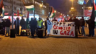 این تصویر مربوط به تجمعی است که در آن تعداد زیادی از افراد با پرچمهای فلسطینی و یک banner بزرگ حضور دارند. روی بنر نوشته شده است: "STOPPT DEN GENOZID IN GAZA" که به معنای "نسلکشی در غزه را متوقف کنید" است. فضای تجمع شهری به نظر میرسد و شب است.