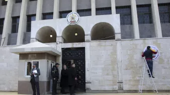 تصویر نشاندهندهٔ یک ساختمان دولتی است که به نظر میرسد در یک کشور عربی قرار دارد. در جلوی ساختمان، شبکهای از ستونها و پنجرهها وجود دارد و بر روی دیوار ورودی، نشان یا علامت مشخصی نصب شده است. چند نفر در جلوی ساختمان ایستادهاند؛ یکی در حال نصب علامت بر روی دیوار و دو نفر دیگر در حال گفتگو به نظر میرسند. همچنین، یک نگهبان در کنار ورودی قابل مشاهده است. زمینهٔ تصویر نیز آسفالت و خطکشی خیابان است.