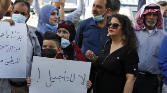 در این تصویر، گروهی از افراد در حال تظاهرات هستند. برخی از آنها ماسک به صورت دارند و تابلوهایی در دست دارند. یکی از تابلوها حاوی جملهای است که به زبان عربی نوشته شده و به معنای "نه به تاخیر!" است. افراد در تصاویر شامل زنان و مردانی با پوششهای مختلف هستند، و برخی از آنها با چهرههای جدی و مصمم به بیان خواستههای خود پرداختهاند. فضای کلی نشاندهندهی اعتراض و تلاش برای بیان نظرات و مطالبات است.