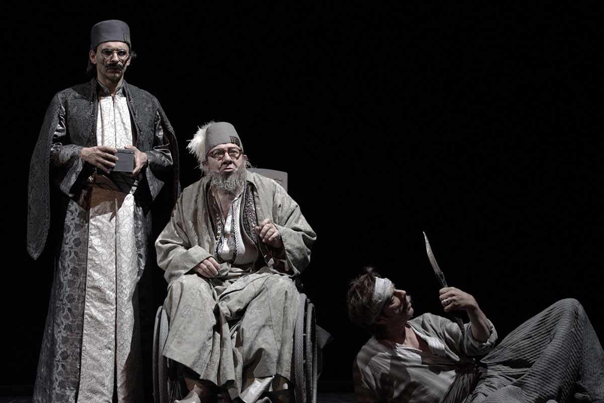 L'image représente une scène de théâtre mettant en avant trois personnages. Deux d'entre eux, habillés de façon traditionnelle et avec des vêtements sombres et ornés, semblent être en position d'autorité ou d'observation. Le troisième personnage est assis dans un fauteuil roulant, avec des vêtements plus clairs et un air pensif ou mélancolique. Il a une longue barbe grise et des lunettes. À côté de lui, un autre personnage, qui paraît être en détresse ou blessé, est au sol, tenant un objet pointu. L'éclairage est sombre, créant une atmosphère dramatique et intense.