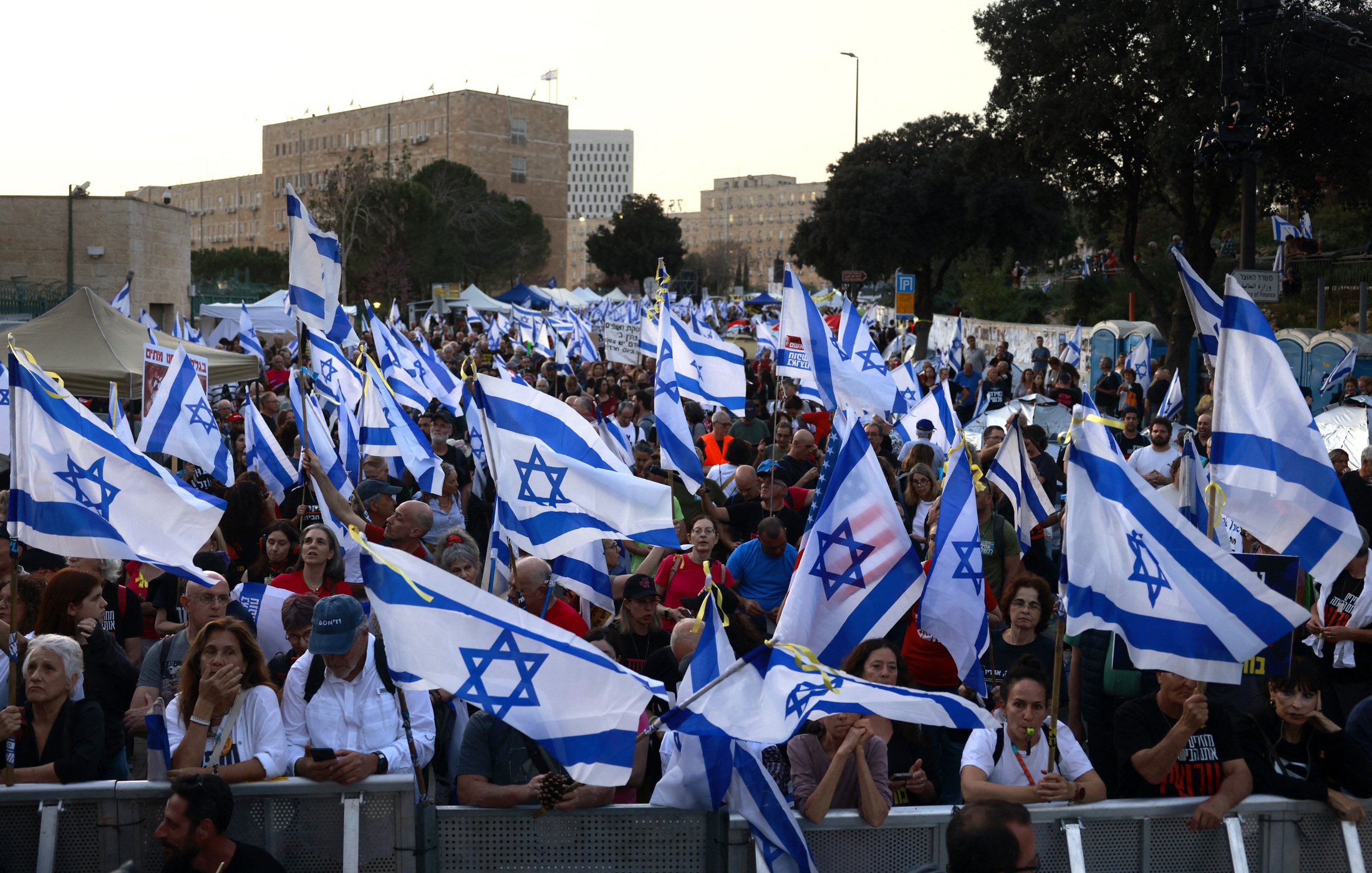 L'image montre une grande foule de personnes rassemblées, brandissant des drapeaux israéliens. Les manifestants semblent réunis pour une occasion particulière, avec une ambiance dynamique. En arrière-plan, on peut apercevoir des bâtiments et des structures urbaines, tandis que des tentes sont installées, peut-être pour fournir des services aux participants. L'ensemble illustre une manifestation significative, avec une forte présence de la couleur bleue et blanche des drapeaux.