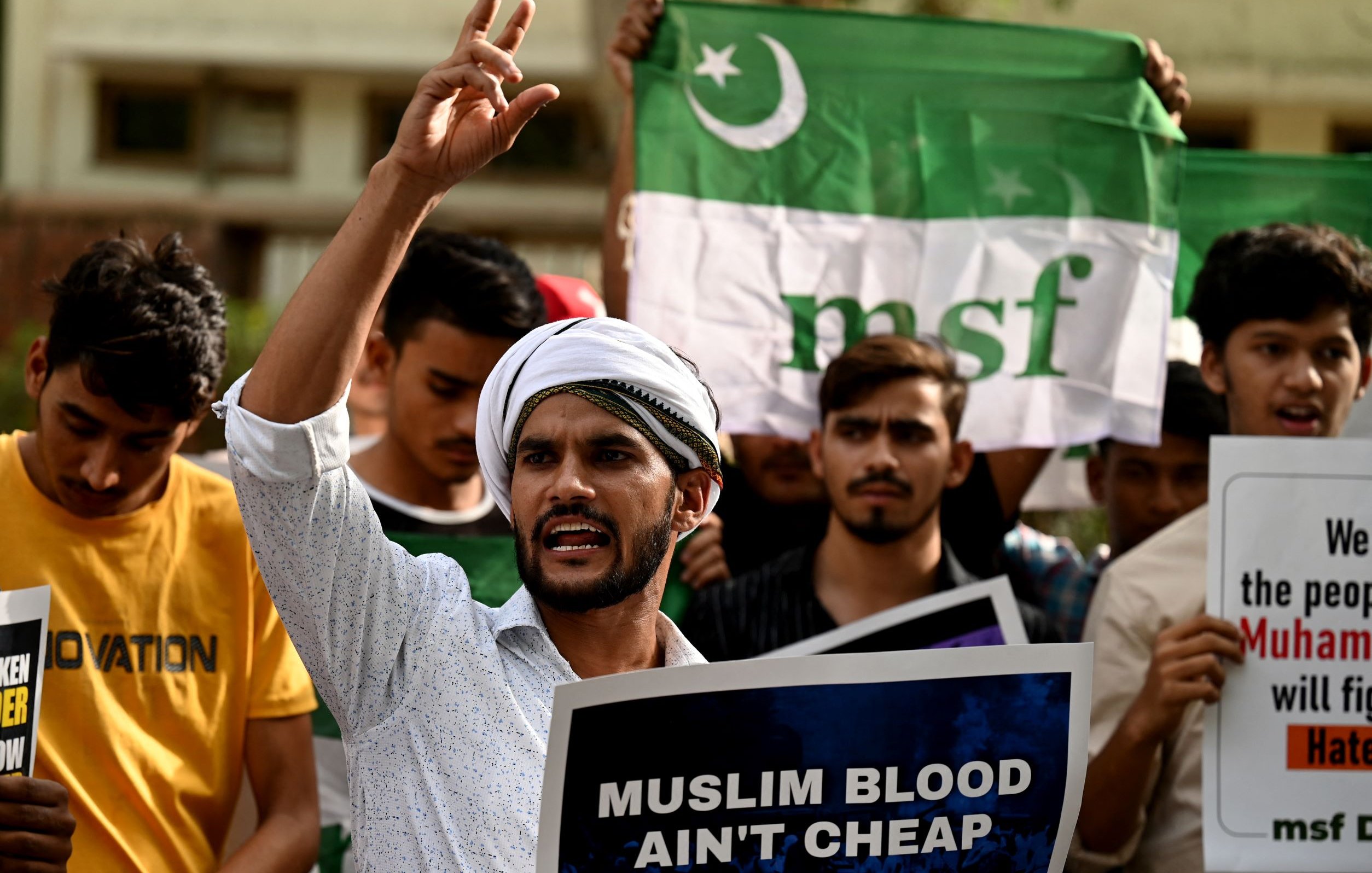L'image montre un groupe de manifestants rassemblés, tenant des pancartes et des drapeaux. Au premier plan, un homme s'exprime avec passion, levant un doigt en l'air. Il porte une chemise claire et un turban. Les autres participants dans le fond semblent également engagés dans la manifestation, brandissant des affiches avec des messages. L'atmosphère semble intense et revendicative.
