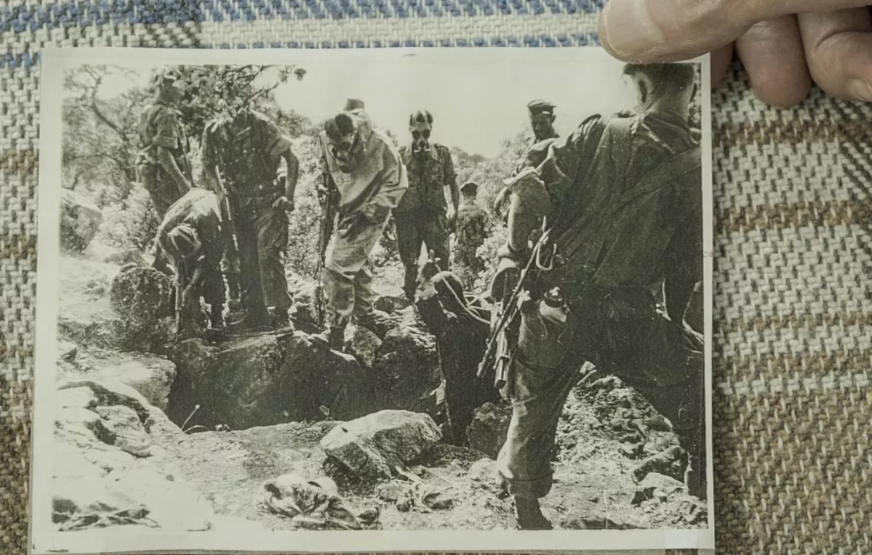 L'image montre un groupe de personnes en uniforme militaire, probablement des soldats, rassemblés autour d'un trou dans le sol. Certains semblent être en train de creuser ou d'examiner le terrain, tandis que d'autres observent. L'ambiance est sérieuse, indicative d'une situation d'urgence ou de conflit. Le tout est en noir et blanc.
