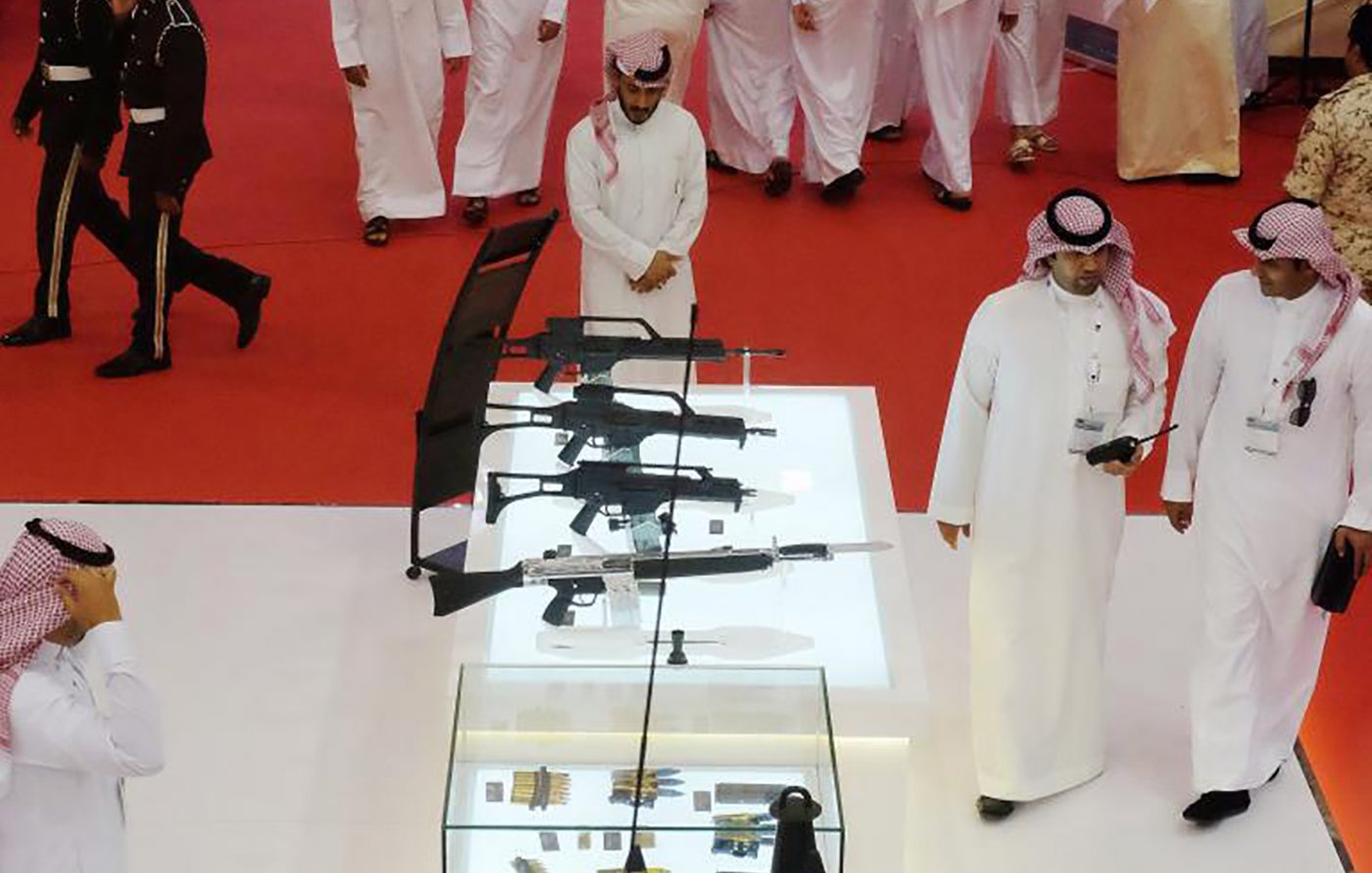 L'image montre un rassemblement où plusieurs personnes, probablement en tenue traditionnelle, se déplacent dans un espace décoré. Au centre, il y a une vitrine en verre contenant diverses armes, mettant en avant des fusils et d'autres équipements. L'ambiance semble formelle et le sol est recouvert d'un tapis rouge. Des membres de la sécurité ou des uniformes militaires peuvent être aperçus en arrière-plan, soulignant l'importance de l'événement.