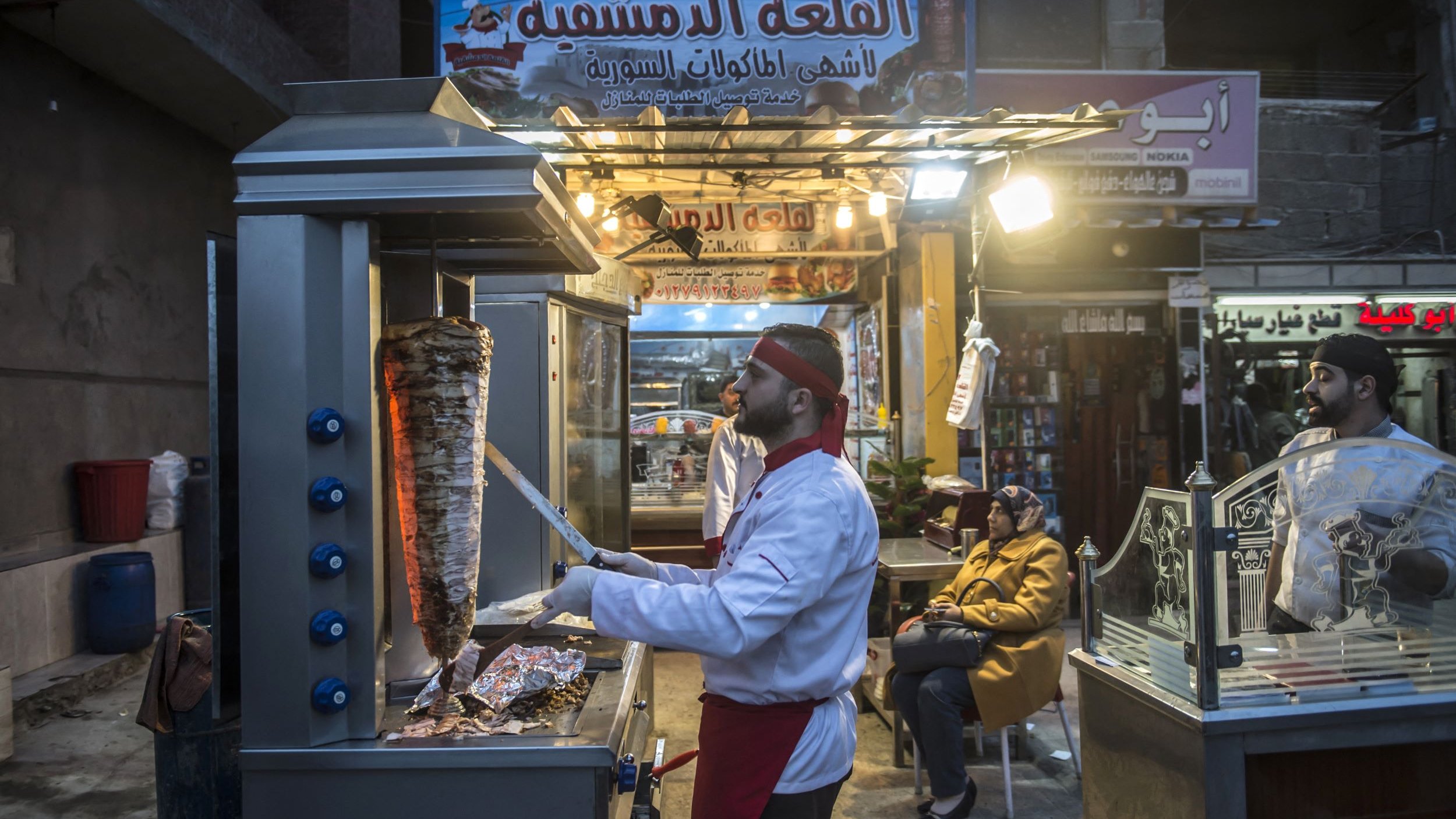 Sur cette image, on peut voir un vendeur de nourriture dans une rue animée, probablement dans un pays du Moyen-Orient. Il est en train de préparer un plat cuisiné, et on peut apercevoir un grand rouleau de viande, typique des stands de kebab ou de shawarma. Le vendeur porte une tenue de chef, composée d'une veste blanche et d'un couvre-chef. À côté de lui, une autre personne semble attendre, assise et observant. L'ambiance est chaleureuse, avec des lumières attirantes au-dessus du stand et des panneaux indiquant le nom du lieu.