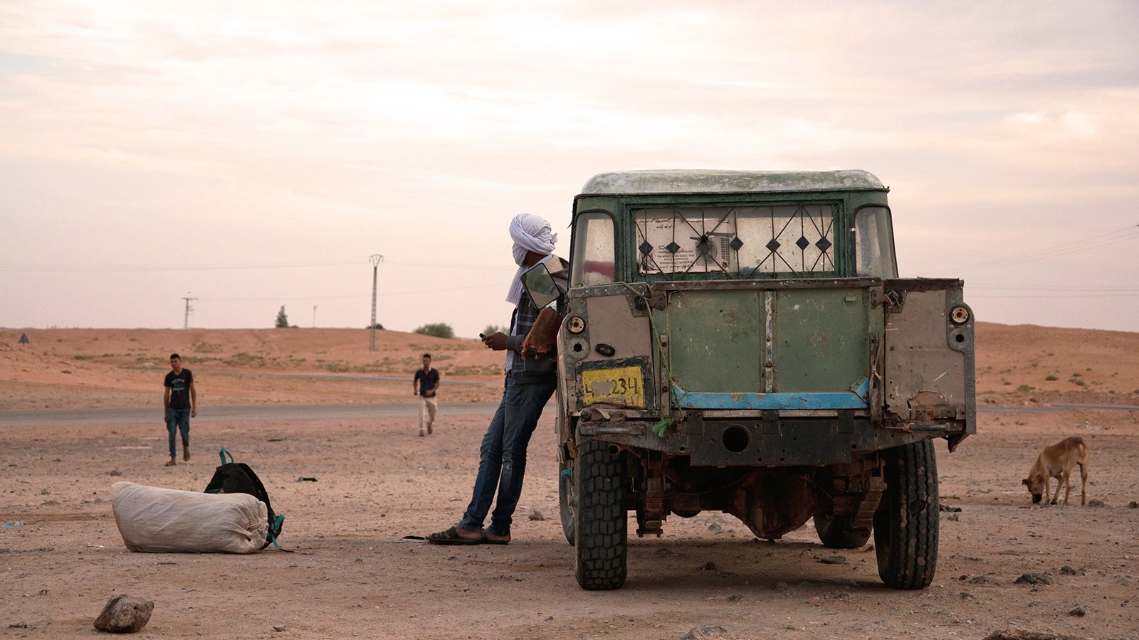 L'image montre une scène en plein air dans un paysage désertique. On peut voir un ancien véhicule utilitaire, avec une carrosserie usée et des fenêtres cassées. Un homme, partiellement visible, se tient à côté du véhicule, portant un turban. En arrière-plan, il y a d'autres personnes, ainsi qu'un chien qui déambule. Le ciel est nuageux, et l'atmosphère semble calme, mais aussi un peu désolée, avec des dunes de sable à l'horizon.