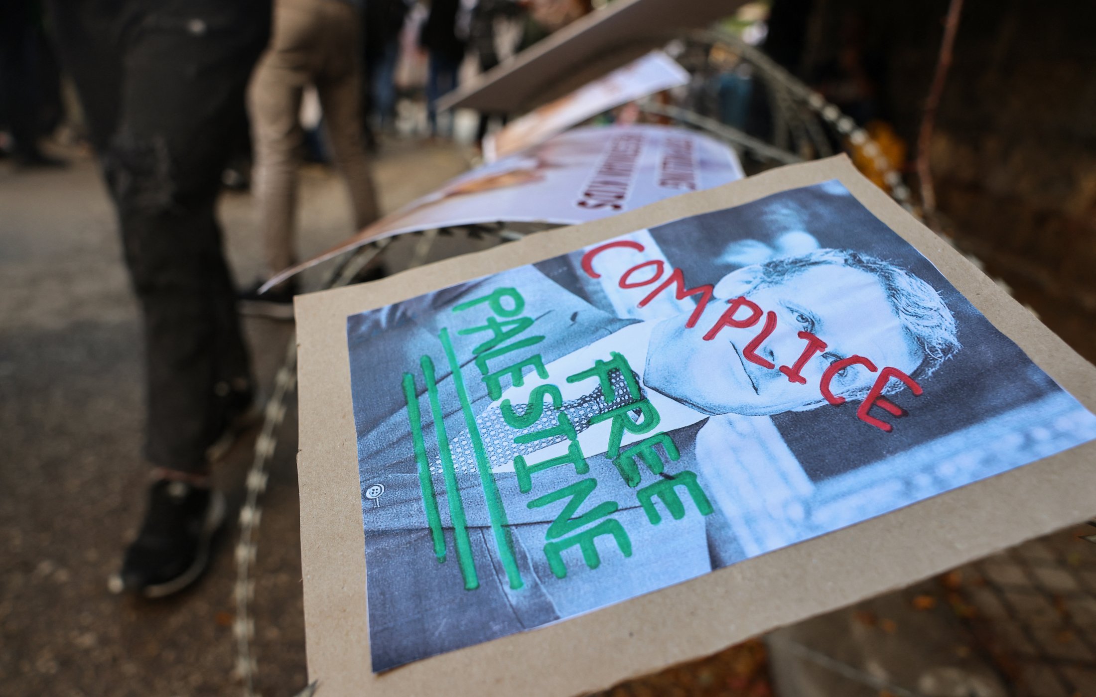 L'image montre des affiches placées sur une barrière, probablement dans le cadre d'une manifestation. L'une des affiches présente un portrait en noir et blanc avec les mots "COMPLICE" et "FREE PALESTINE" écrits en rouge et vert. L'environnement semble être celui d'une manifestation ou d'un rassemblement, avec plusieurs personnes en arrière-plan.