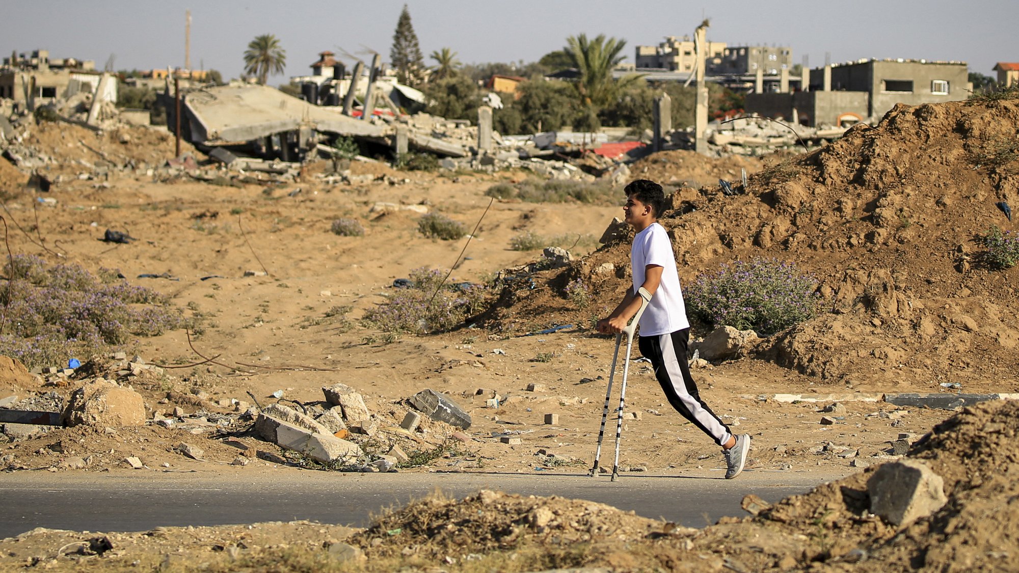 L'image montre un jeune garçon marchant avec des béquilles dans un paysage de destruction. En arrière-plan, on peut voir des bâtiments en ruines et des débris éparpillés, témoignant d'un environnement dévasté. Le sol est recouvert de terre et de petites herbes, créant un contraste avec les restes des structures. L'atmosphère semble sombre et tragique, soulignant la situation difficile dans laquelle se trouve l'enfant.