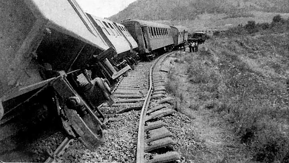 L'image montre un accident de train. Plusieurs wagons sont déraillés, penchés sur le côté, tandis que les rails semblent déformés. En arrière-plan, on aperçoit des montagnes et des personnes qui semblent inspecter les lieux. L'atmosphère est celle d'un événement tragique, évoquant une situation d'urgence.