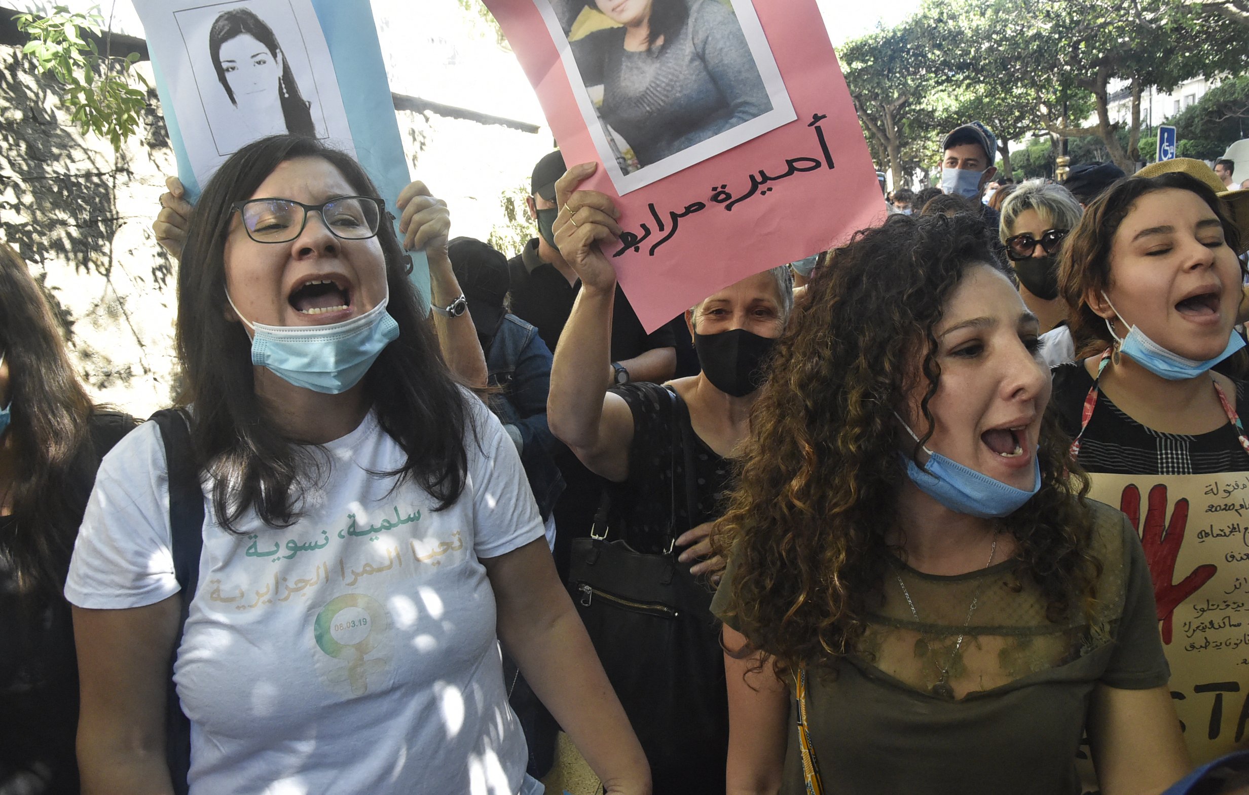L'image montre un groupe de manifestants lors d'une manifestation. Plusieurs personnes tiennent des pancartes avec des photographies et des slogans. Les participants semblent exprimés des émotions fortes, comme la colère ou la détermination, en scandant des slogans. Certaines personnes portent des masques de protection, tandis que l'environnement semble urbain avec des arbres et des bâtiments en arrière-plan.