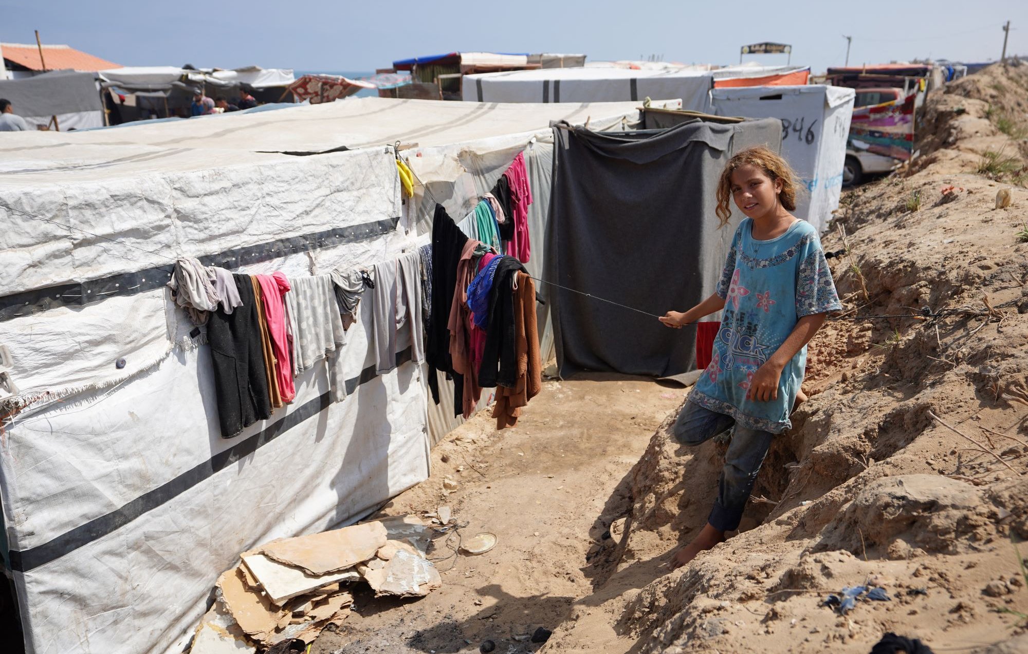 L'image montre un camp de tentes ou de structures temporaires. À gauche, des vêtements sont accrochés à une corde à linge, tandis qu'une jeune fille se trouve au premier plan, tenant une sorte de bâton. Elle porte une tunique et semble se tenir sur un chemin étroit entre les tentes. Le décor est marqué par des bords de sol en terre et la structure du camp qui se développe à l'arrière-plan. L'atmosphère semble celle d'un environnement difficile, lié à des conditions de vie précaires.