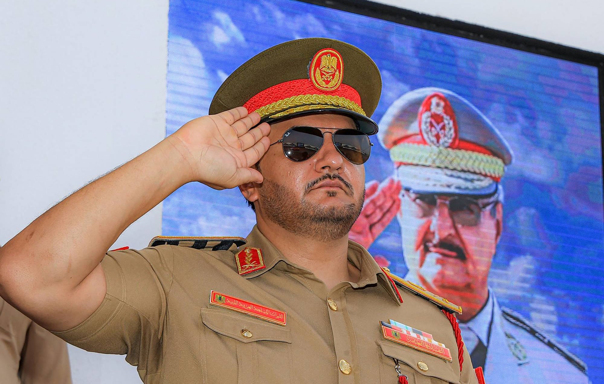 Un homme en uniforme militaire, lunettes noires, salue devant un écran.