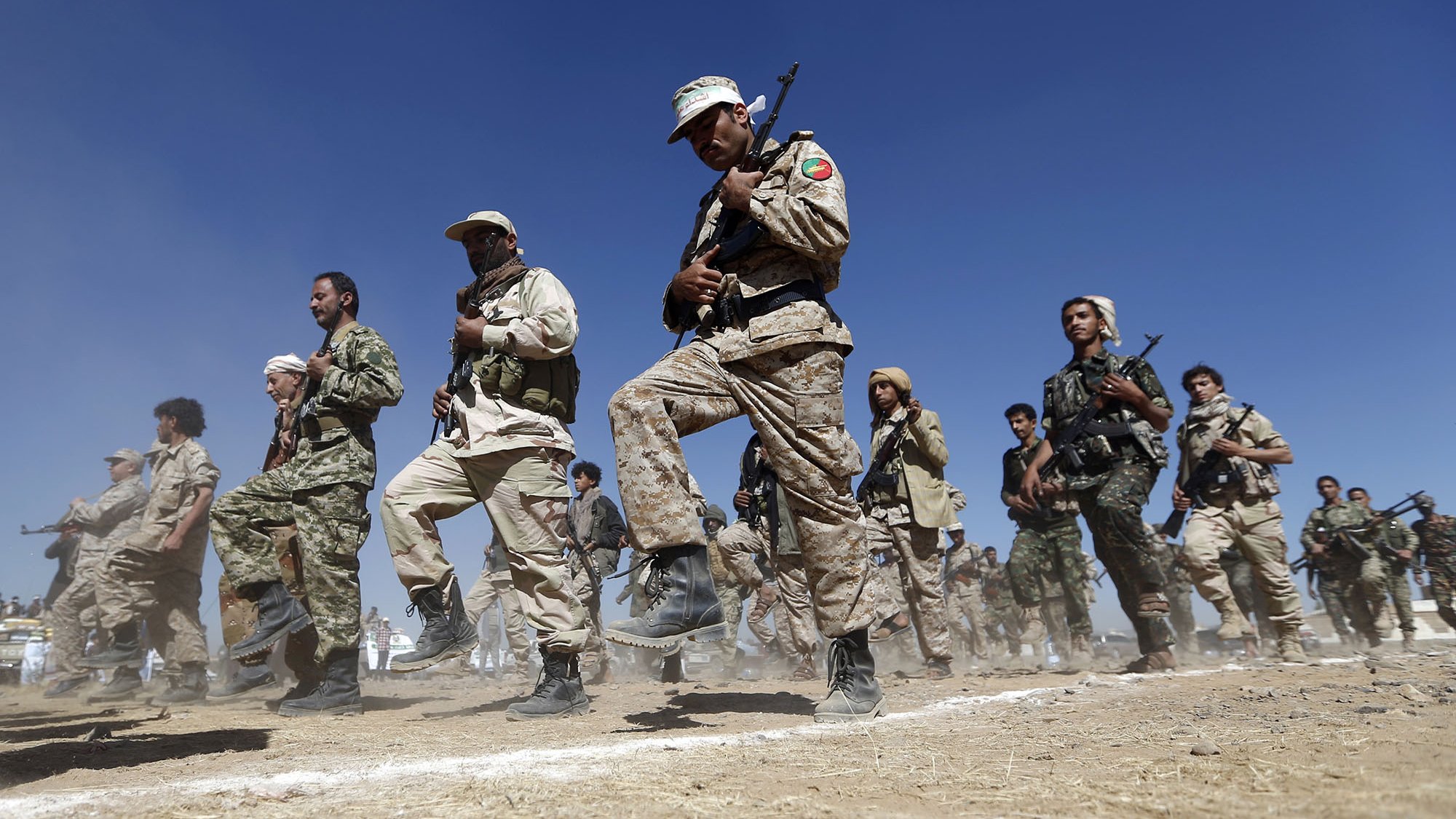 L'image montre un groupe de personnes en uniforme militaire en train de marcher ou de s'entraîner. Ils semblent être réunis en formation, tenant des fusils. Le terrain est poussiéreux, et on peut apercevoir un ciel bleu en arrière-plan. L'ambiance semble sérieuse et déterminée, suggérant une activité militaire ou un exercice de préparation.
