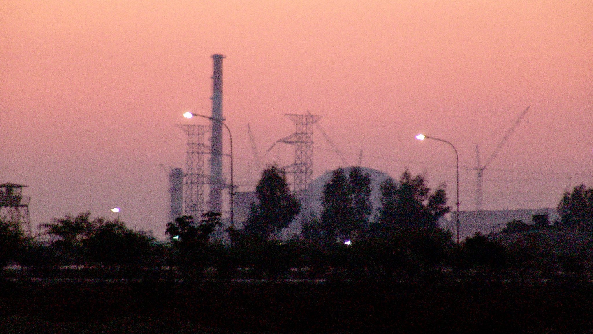 L'image montre une scène de crépuscule avec un ciel teinté de rose et d'orange. Au premier plan, on peut voir des arbres ainsi que des lampadaires allumés. En arrière-plan, se dressent des structures industrielles, notamment des cheminées et des pylônes électriques, ainsi que des grues de construction, ce qui suggère une activité industrielle en cours. L'atmosphère est calme et paisible, malgré la présence des installations.