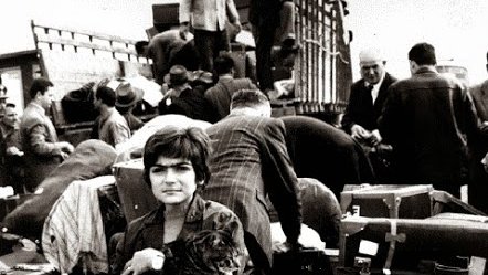 L'image montre une scène de personnes en situation de déplacement. Au premier plan, une jeune fille tient un animal, probablement un chat, dans ses bras, et elle est entourée de nombreux bagages, valises et paquets. En arrière-plan, on aperçoit d'autres personnes, certains sont en train de charger ou de décharger des objets d'un véhicule. L'atmosphère est chargée, et l'image semble capturer un moment de transition ou d'exode, évoquant des sentiments de nostalgie et de désespoir. Les tonalités en noir et blanc ajoutent une dimension dramatique à la scène.