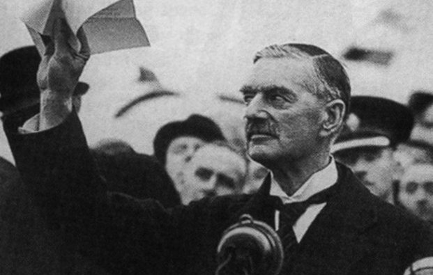 L'image montre un homme en costume, se tenant devant un microphone avec une expression grave. Il lève un ou plusieurs documents au-dessus de sa tête, visiblement en train de s'adresser à une foule. En arrière-plan, on peut voir des personnes assemblées, portant des uniformes et écoutant attentivement. L'atmosphère semble être celle d'un discours important, probablement lié à des événements politiques.