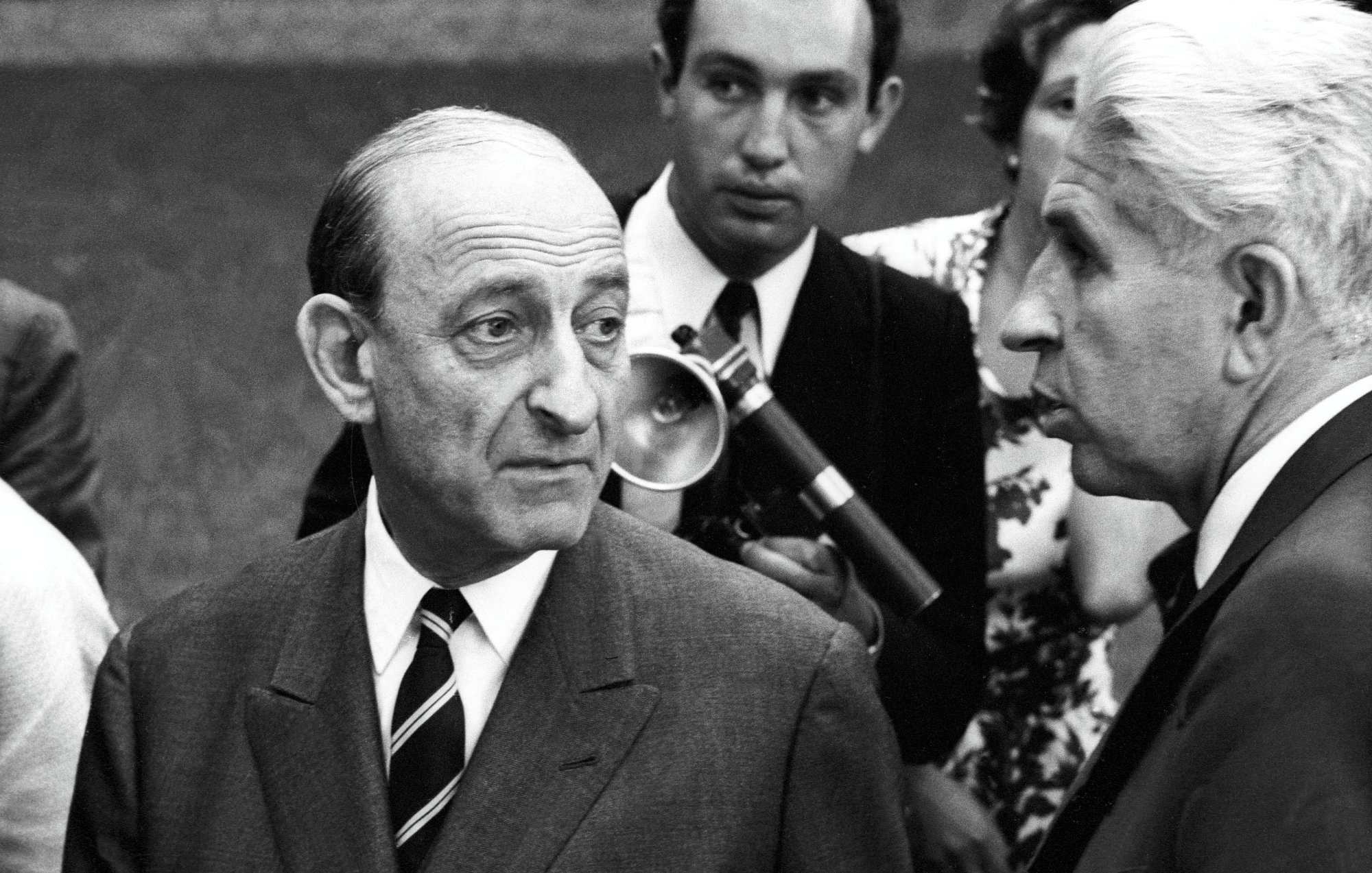 Deux hommes en costume, discutant sérieusement, entourés d'autres personnes. Époque rétro.