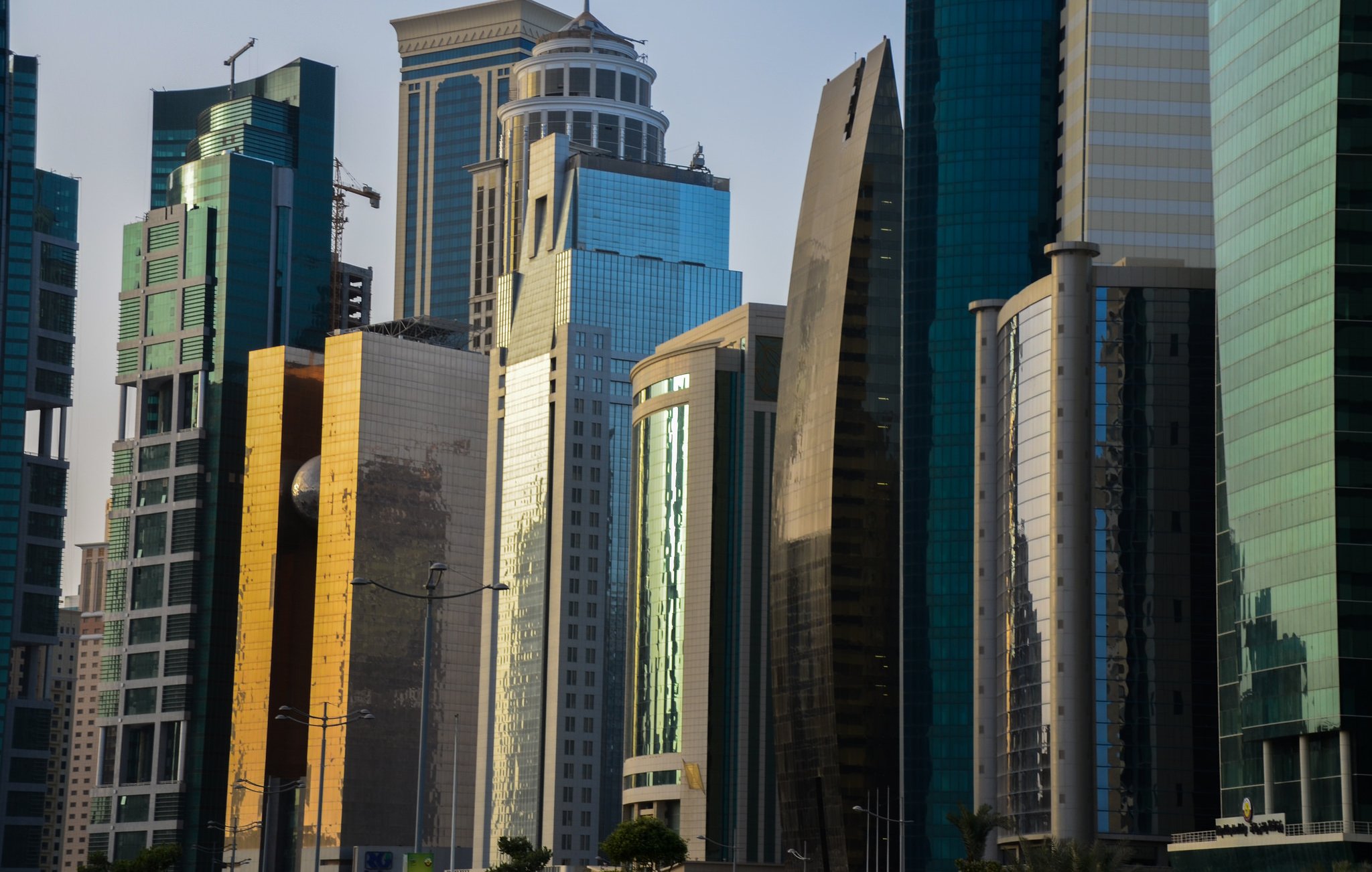 L'image montre une vue urbaine moderne avec plusieurs gratte-ciels aux façades brillantes et réfléchissantes. Les bâtiments sont de différentes formes et tailles, avec des matériaux en verre et en métal qui scintillent sous la lumière. On peut remarquer un mélange d'architectures contemporaines, certaines avec des courbes élégantes et d'autres plus angulaires. Le paysage urbain reflète un environnement dynamique et en pleine croissance, typique des grandes villes modernes.