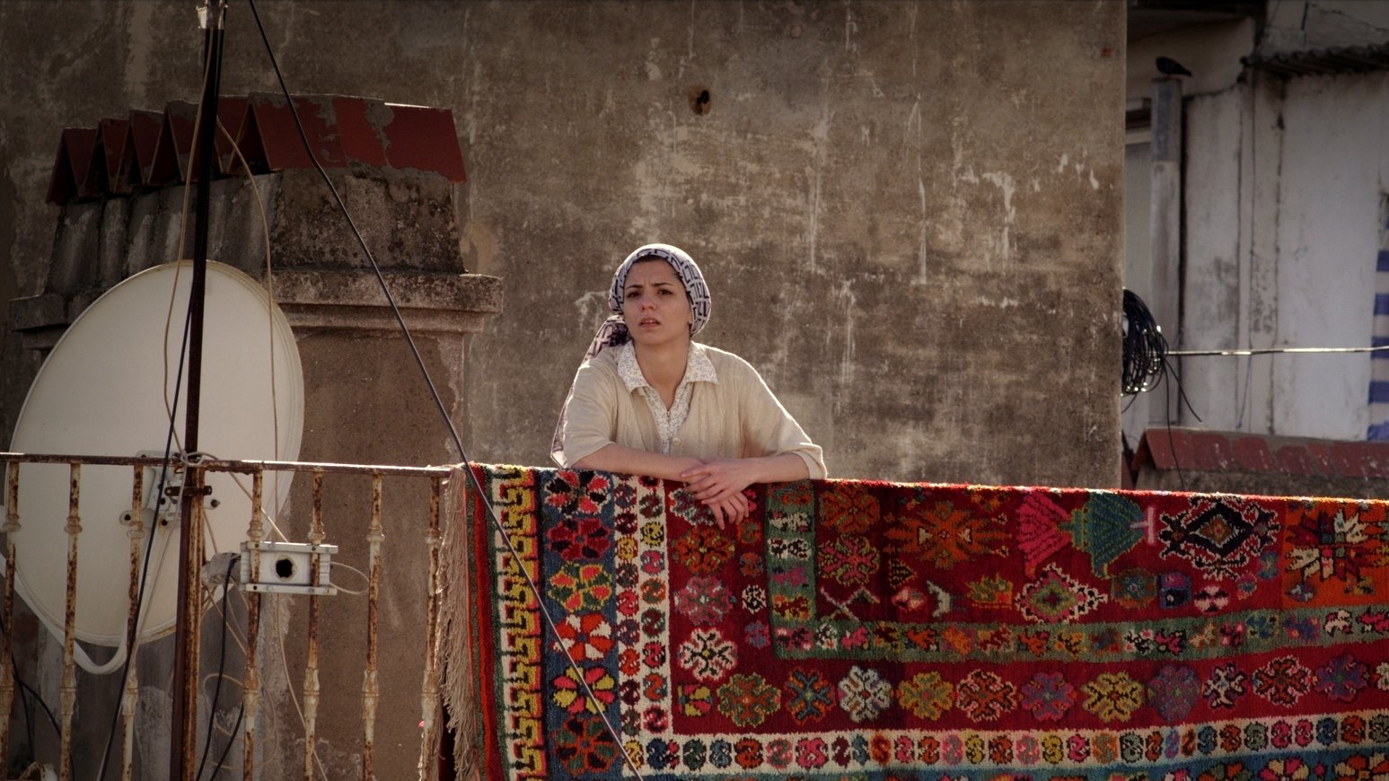 L'image montre une femme se tenant sur un balcon, regardant au loin. Elle porte un haut clair et un foulard sur la tête. Derrière elle, un grand tapis coloré est accrochée à la balustrade, avec des motifs détaillés et vibrants. En arrière-plan, on aperçoit un mur et une antenne satellite, ce qui suggère un environnement urbain. L'éclairage donne une atmosphère chaleureuse à la scène.