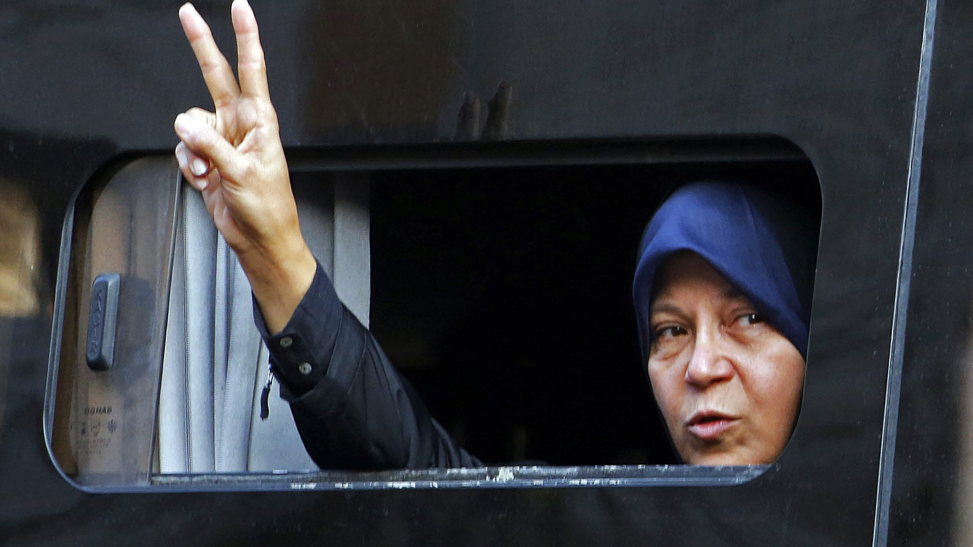 L'image montre une femme qui fait un signe de paix avec sa main, en levant deux doigts. Elle est visible à travers une fenêtre légèrement ouverte d'un véhicule. La femme porte un hijab. Son expression semble déterminée et engagée. Le fond est sombre, ce qui met en avant son geste et son visage.