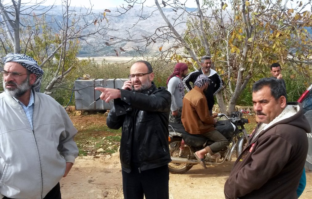 L'image montre un groupe d'hommes dans un environnement rural, probablement en plein air avec des arbres autour. Un homme, habillé avec une veste noire, est au centre, en train de parler au téléphone et de pointer du doigt dans une direction. D'autres hommes sont dispersés autour de lui, certains se tenant debout, d'autres assis sur une moto. L'arrière-plan montre des collines ou des montagnes, suggérant une zone naturelle. L'atmosphère semble détendue mais attentive, comme si les hommes discutaient d'un sujet important.