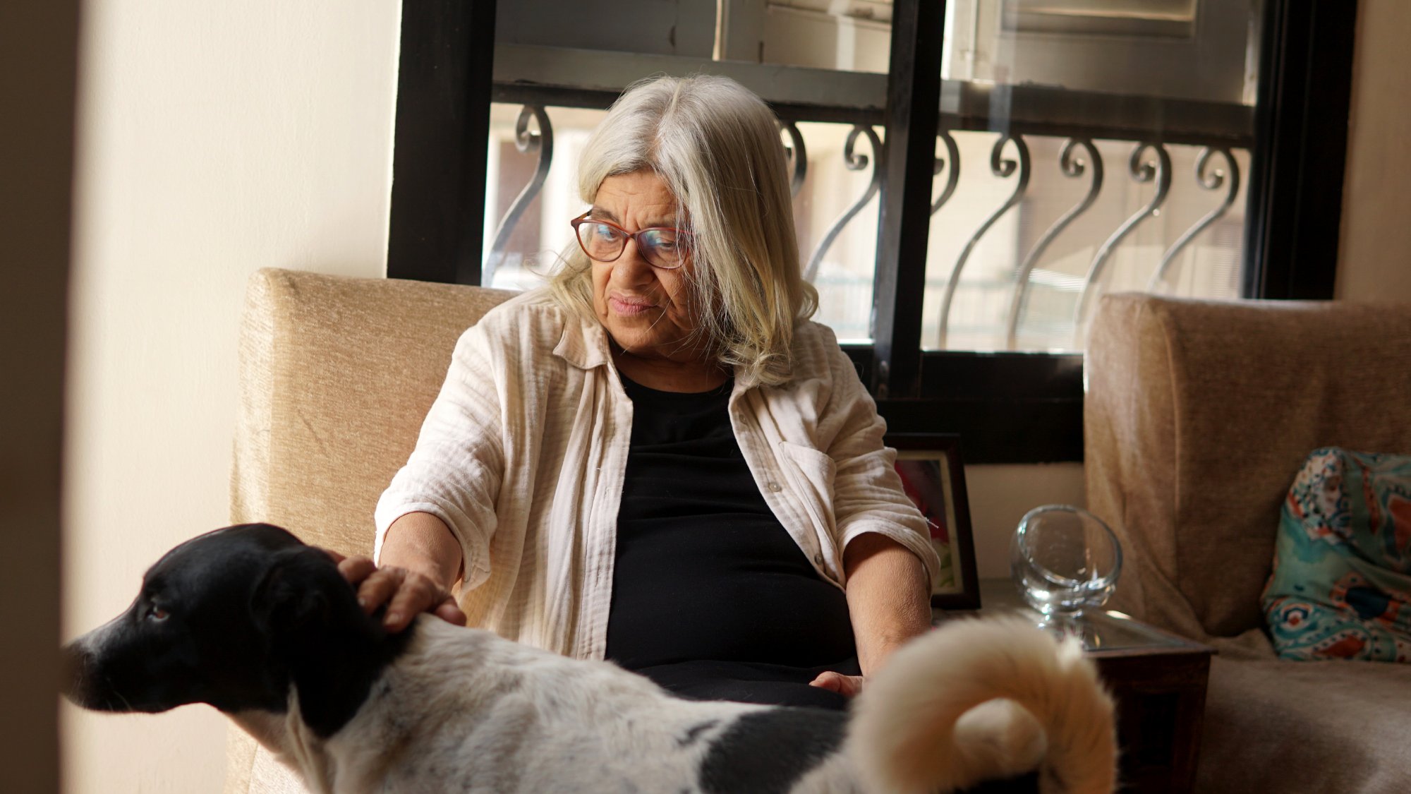 L'image montre une femme âgée avec de longs cheveux gris assise dans un fauteuil. Elle porte des lunettes et regarde avec tendresse un chien noir et blanc qui se trouve près d'elle. La lumière entre par la fenêtre, créant une atmosphère chaleureuse et douce dans la pièce. Des éléments de décoration sont visibles en arrière-plan, ce qui ajoute à l'ambiance accueillante de l'endroit.