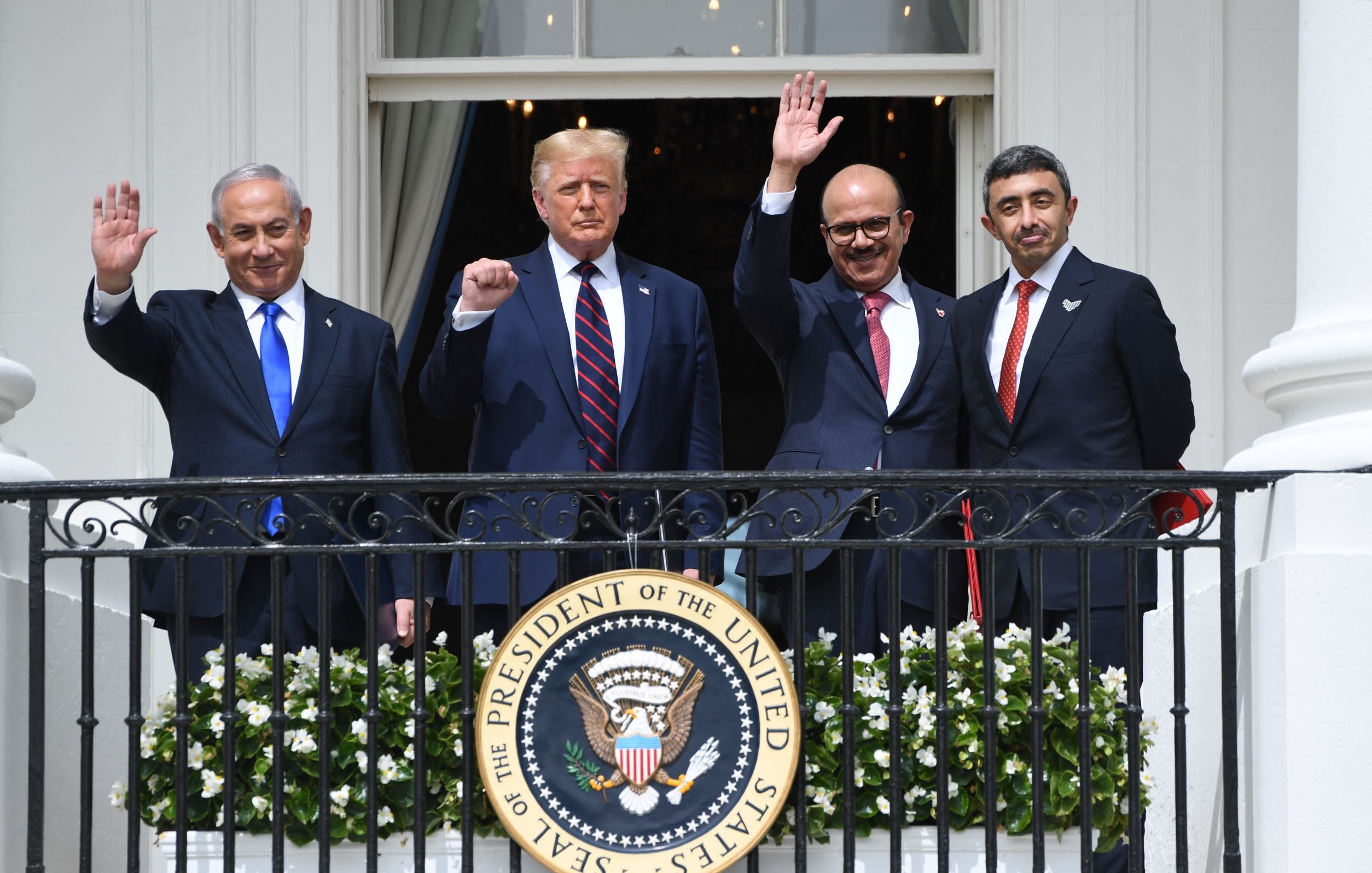 L'image montre quatre hommes debout sur un balcon de la Maison Blanche, levant la main en signe de salutation. Ils semblent être en train de célébrer un moment important, probablement lié à des événements diplomatiques. L'un des hommes porte un costume sombre avec une cravate rouge, tandis que les autres affichent également des tenues formelles. En arrière-plan, on peut voir un environnement architectural caractéristique de la Maison Blanche, et un grand sceau du Président des États-Unis est en évidence sur la balustrade.