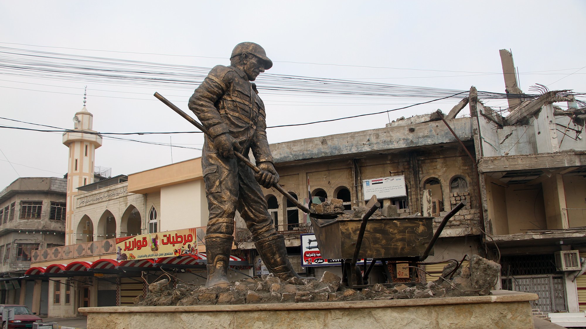 L'image montre une statue monumentale représentant un travailleur en train de creuser, symbolisant probablement la reconstruction ou le labeur dans un contexte difficile. En arrière-plan, on distingue des bâtiments partiellement détruits et des infrastructures urbaines. La scène évoque une atmosphère de résilience et de reconstruction dans une zone touchée par des conflits. La statue est installée sur un socle en pierre, ajoutant à son importance et à son impact visuel.