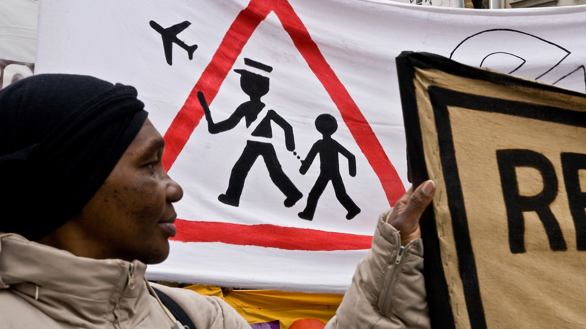 L'image montre une manifestation avec une femme tenant une pancarte. En arrière-plan, il y a une grande bannière avec un symbole d'avertissement en forme de triangle, représentant un adulte marchant avec un enfant. Ce symbole est accompagné d'illustrations d'avions en haut, suggérant une thématique liée au transport ou à la sécurité. La femme porte un manteau clair et un foulard noir, et son expression pourrait refléter l'engagement dans la cause qu'elle soutient.