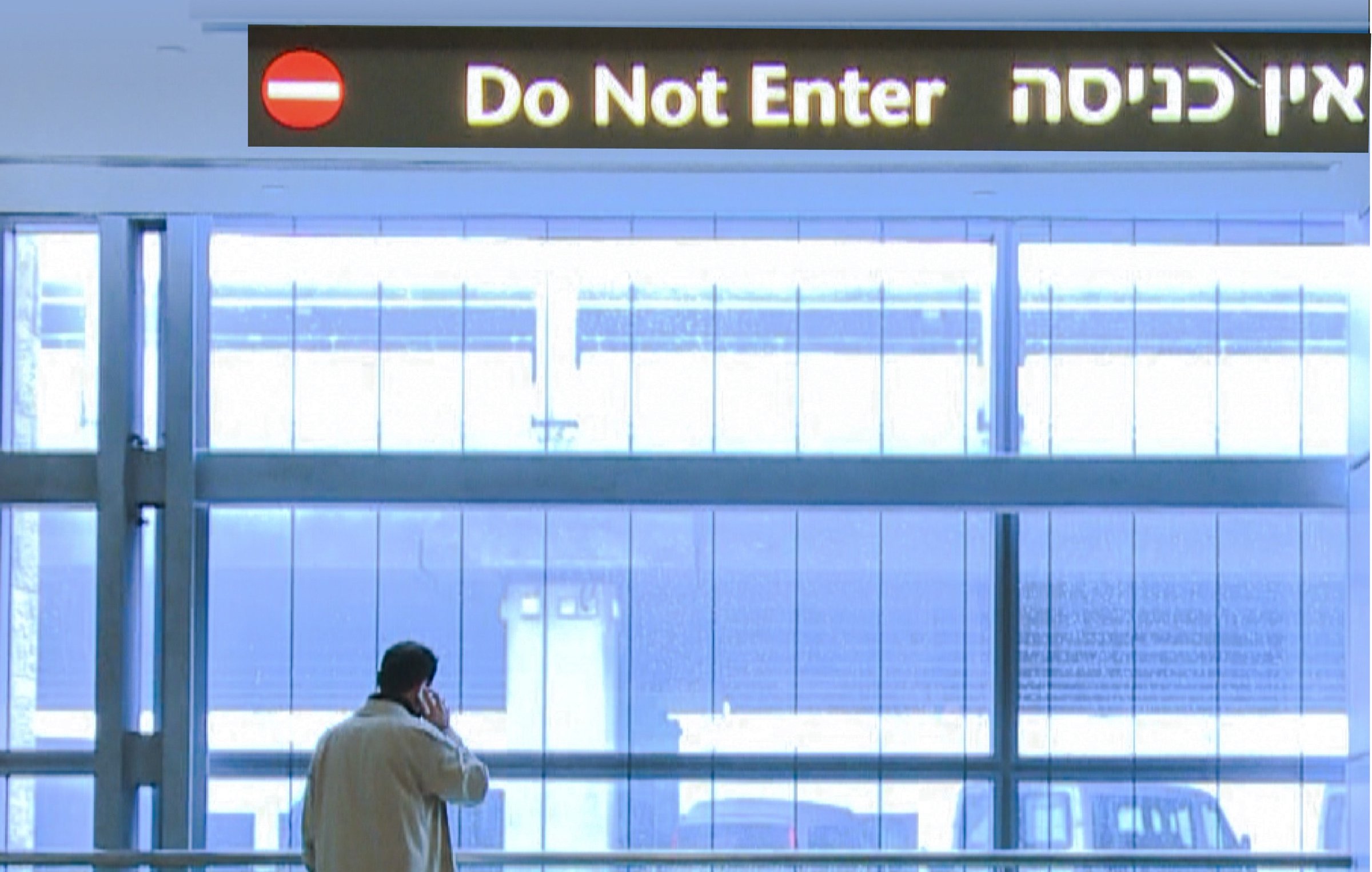 L'image montre un homme debout près d'une grande fenêtre dans un espace intérieur moderne. Il porte un long manteau clair et semble être en train de parler au téléphone. Au-dessus de lui, il y a une signalisation en trois langues, indiquant "Do Not Enter" en anglais et une phrase en hébreu, ainsi qu'un panneau rouge avec un symbole d'interdiction. L'espace est bien éclairé, avec des murs en verre et une ambiance assez minimaliste.