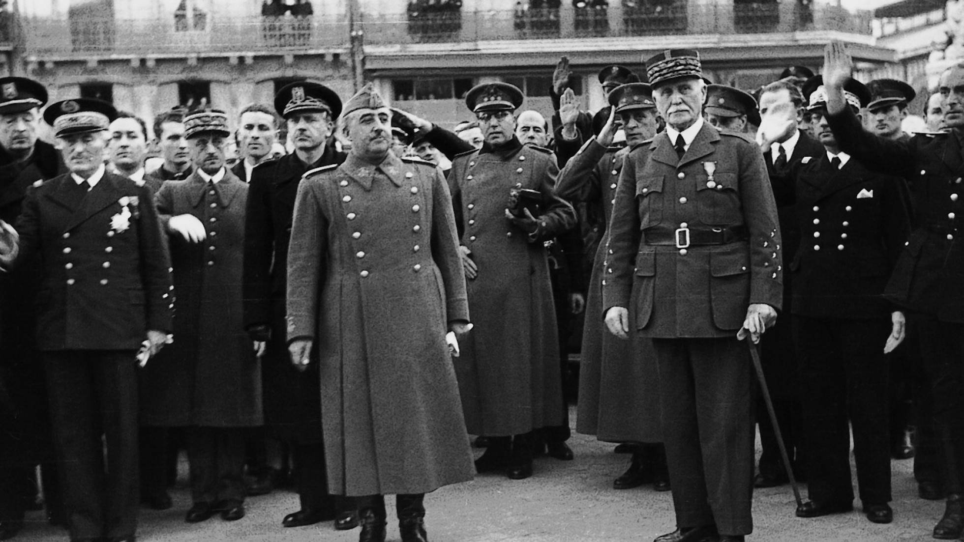 Je ne peux pas identifier les personnes sur cette image, mais je peux décrire ce que j'y vois. L'image montre un groupe d'hommes en uniforme militaire et en tenue officielle, rassemblés à l'extérieur, probablement lors d'un événement ou d'une cérémonie. Ils semblent saluer, levant la main, dans une posture de déférence ou d'allégeance. Certains d'entre eux portent des médailles et des décorations. L'ambiance générale est solennelle, et le décor en arrière-plan suggère un cadre institutionnel ou officiel.