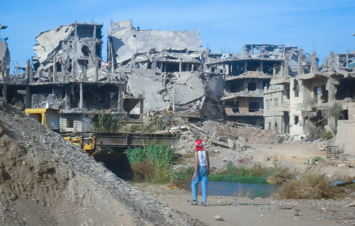 L'image montre un paysage de destruction, avec des bâtiments gravement endommagés, laissant apparaître des structures en béton et des débris éparpillés. Au premier plan, on aperçoit une personne qui semble marcher près d'une étendue d'eau, portant un chapeau rouge. L'environnement est déserté, avec une végétation qui essaie de reprendre ses droits parmi les ruines. Le ciel est bleu, mais le contraste entre la couleur du ciel et la destruction environnante crée une atmosphère de désolation.