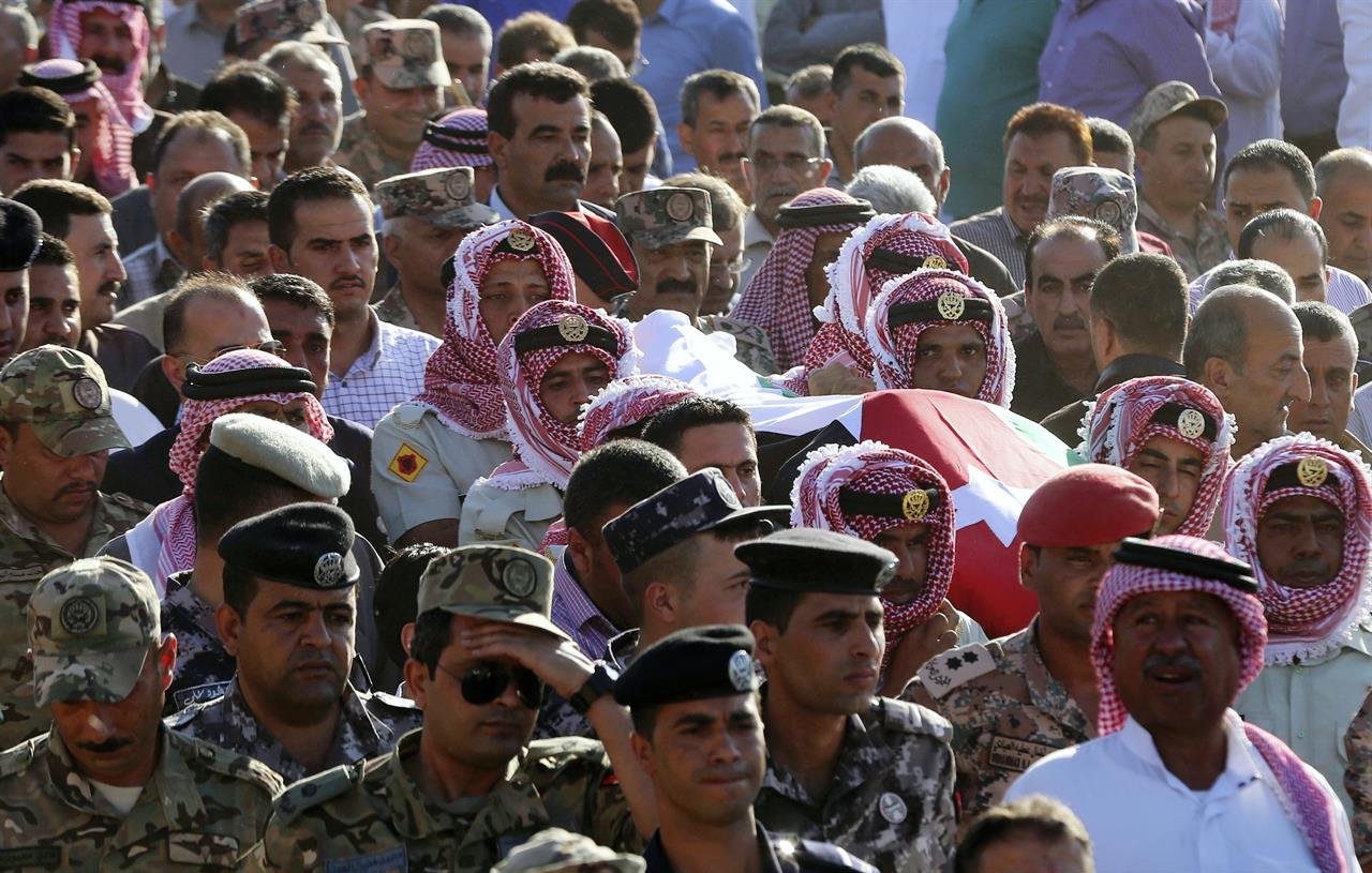 L'image montre une grande foule rassemblée. Au premier plan, des hommes portant des keffiehs (un type de couvre-chef traditionnel) entourent un cercueil recouvert d'un drapeau. Les participants semblent montrer leurs respects, et on peut également voir des militaires en uniforme dans la foule. L'atmosphère semble solennelle, indiquer une cérémonie ou des funérailles d'un individu important.