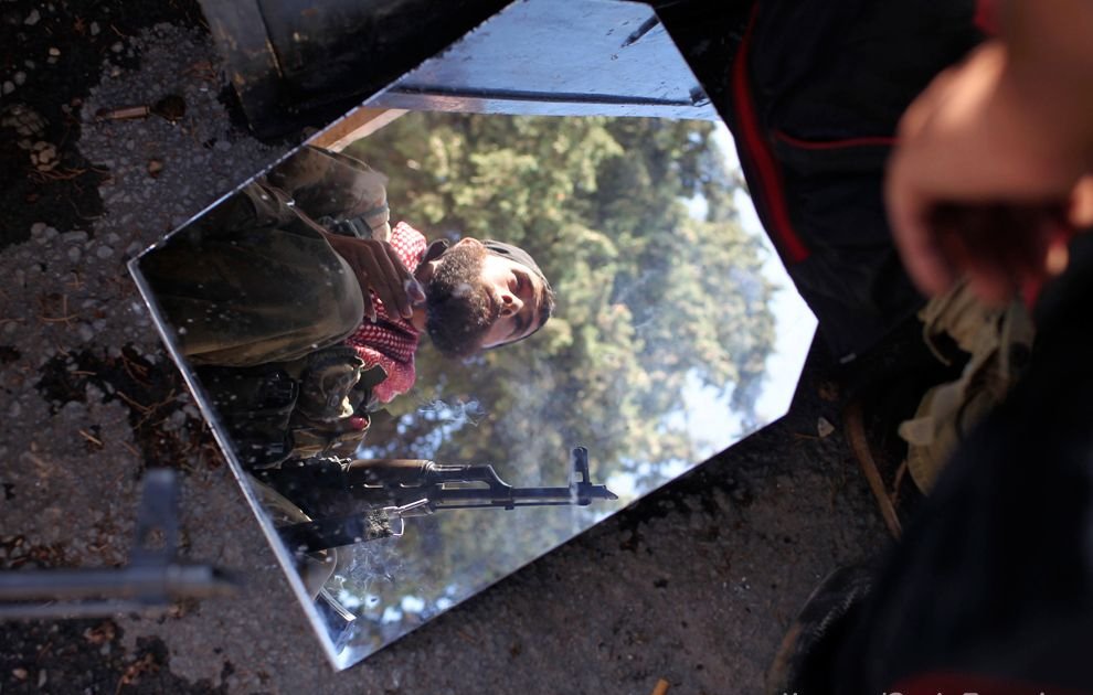 L'image montre un miroir fragmenté reposant sur le sol, reflétant l'image d'un homme en uniforme avec un fusil. À l'arrière-plan, on voit des arbres, suggérant un environnement naturel ou une zone de conflit. Le reflet donne une perspective intrigante sur la scène. L'homme semble vigilant, peut-être en attente ou en préparation pour une opération.