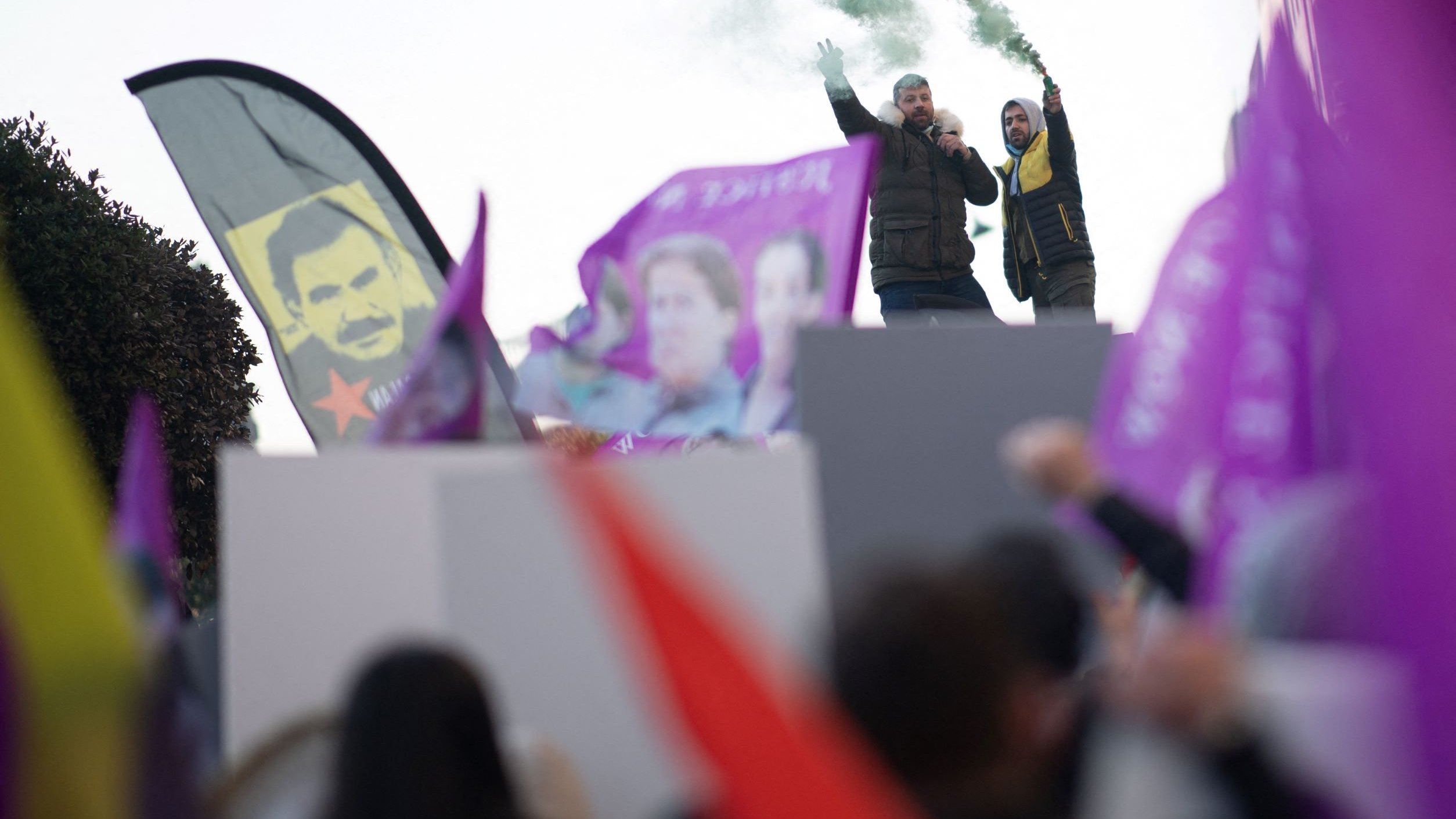 L'image montre une scène de manifestation. Des personnes brandissent des drapeaux et des bannières colorées, principalement en violet et rouge. Au premier plan, on aperçoit deux personnes sur une estrade, l'un d'eux tenant un mégaphone et levant les bras, tandis que l'autre semble encourager la foule. De la fumée verte s'échappe, ajoutant une atmosphère dynamique à l'événement. En arrière-plan, on distingue des portraits, probablement de figures politiques ou symboliques. L'ensemble dégage une ambiance de mobilisation et de passion.